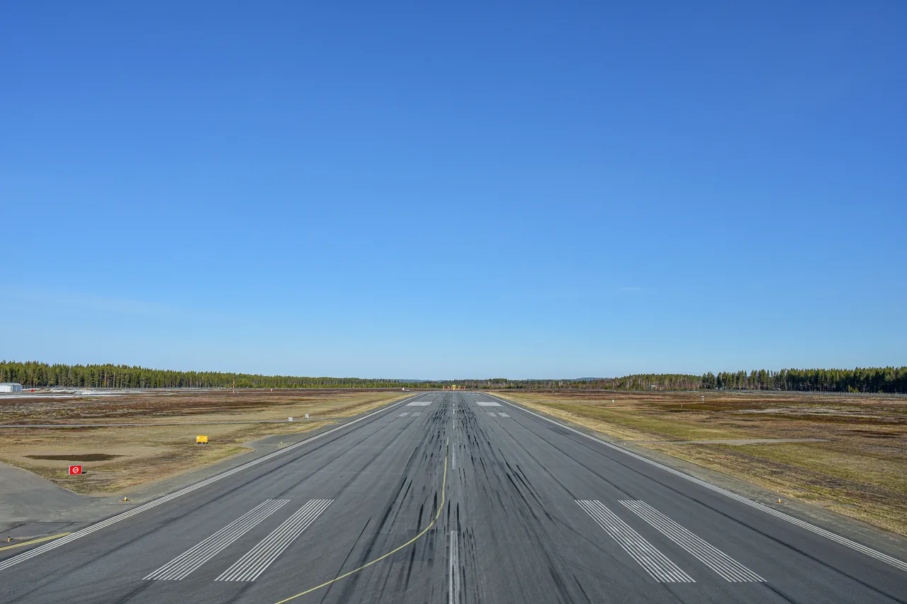 A real asphalt runway with white markings and tire wear, stretching toward a tree-lined horizon under a clear blue sky.